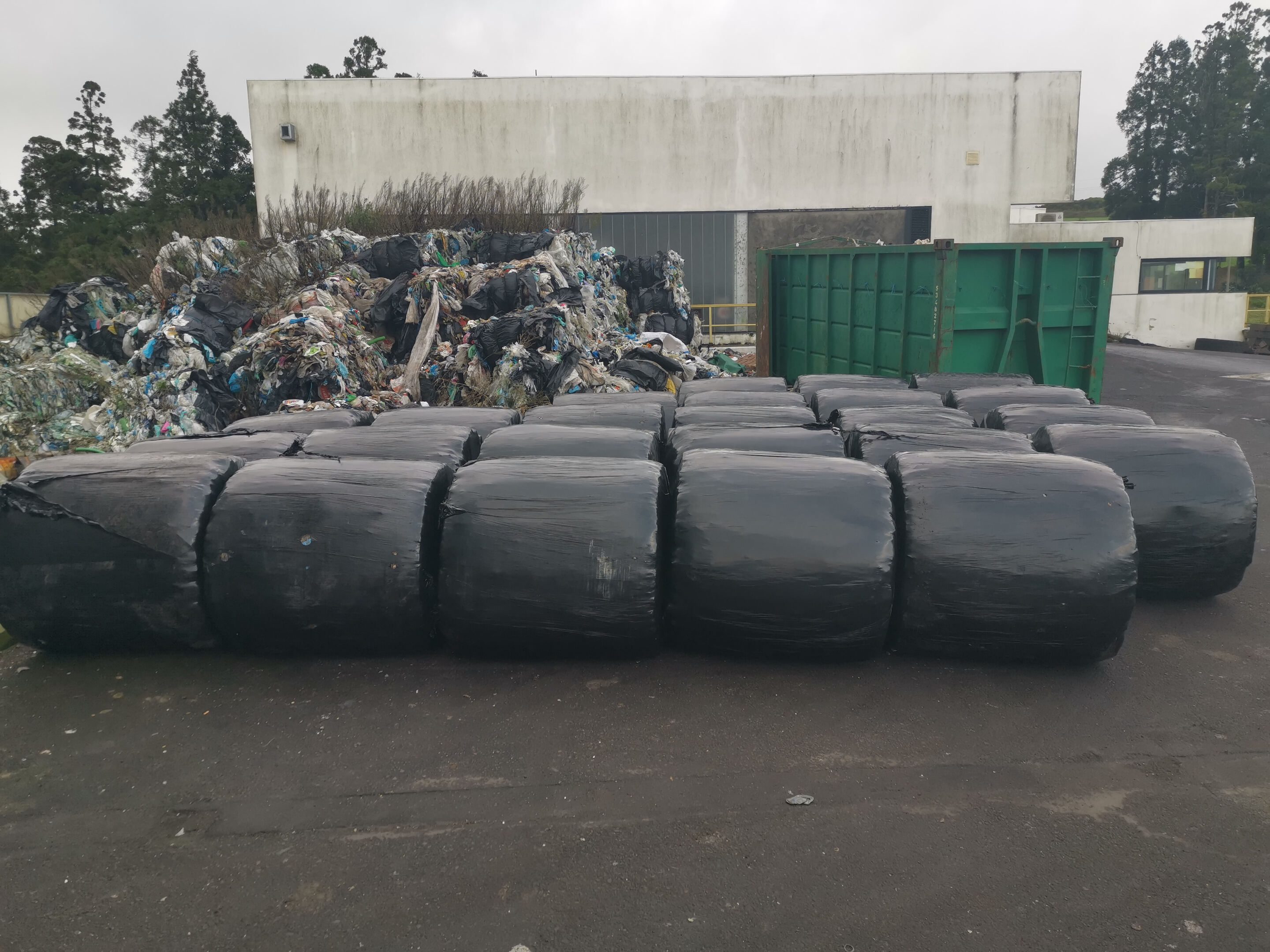 Black round bales lying on their round side with a heap of colored landfill material in the background.