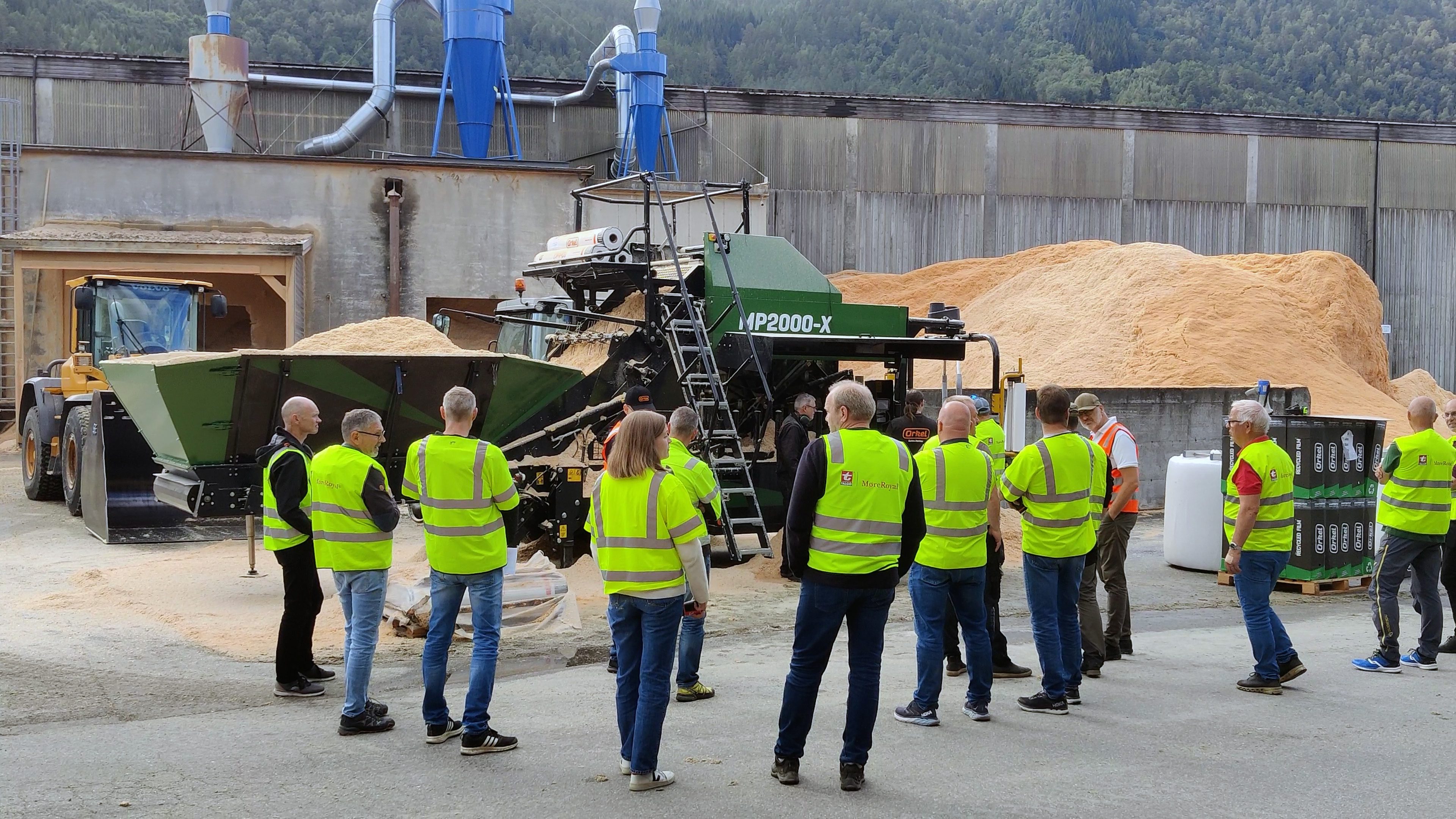 Eine Menschenmenge in gelben Sicherheitswesten steht vor einem MP2000-X, der Holzspäne zu Ballen presst, mit einem großen Haufen Holzspäne im Hintergrund.