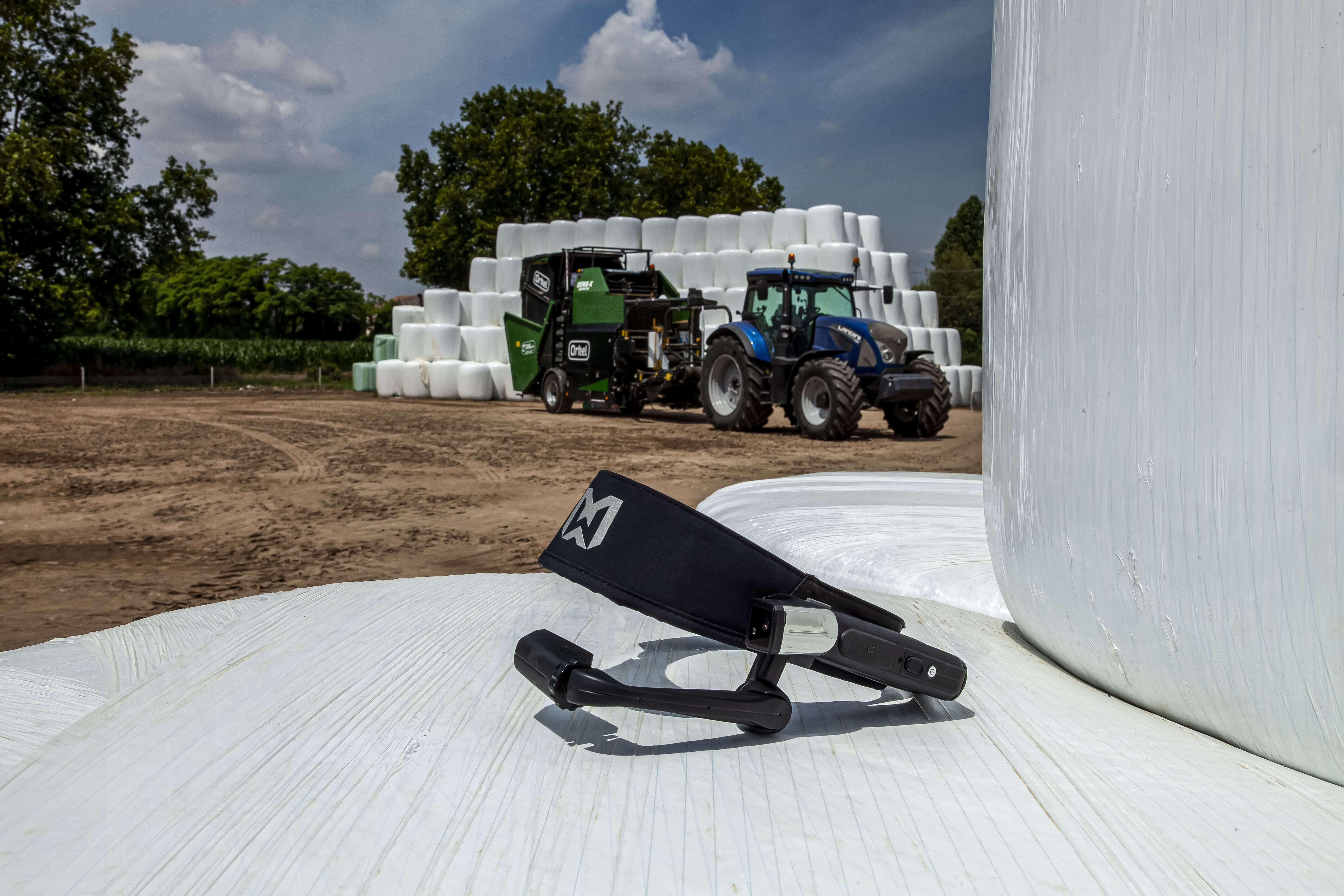 Plano aproximado de um dispositivo RealWear com um compactador Orkel e fardos redondos brancos em segundo plano