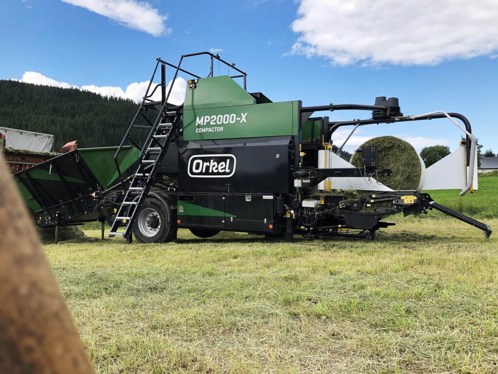 Um compactador Orkel verde com um fardo redondo na mesa de envolvimento, parado num campo com tempo ensolarado, enfardando erva cortada em pequenos pedaços