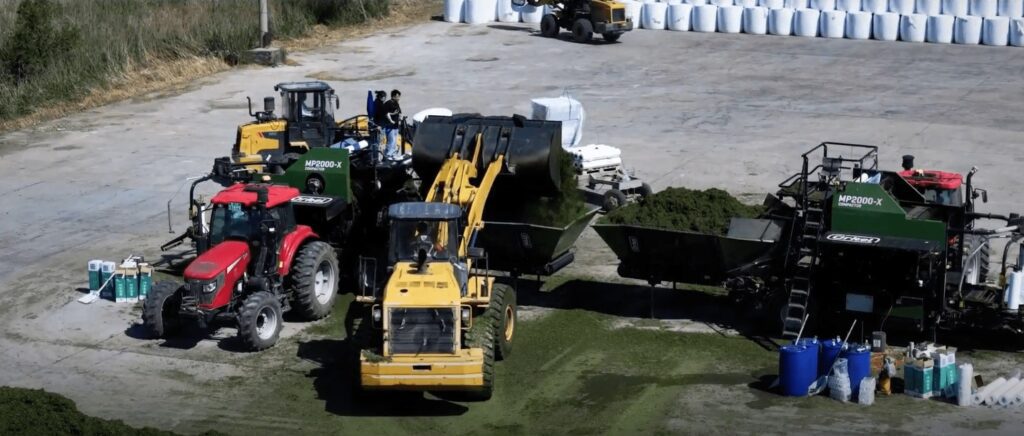 Zwei landwirtschaftliche Ballenpressen pressen Ballen auf einer großen Betonfläche mit Rundballen im Hintergrund. Ein Frontlader kippt Material in die Futtertrichter.