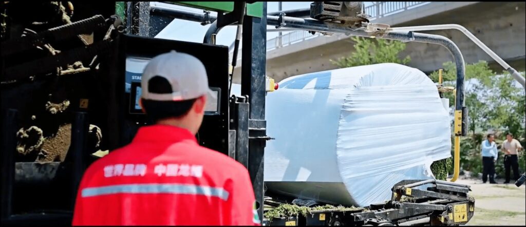 Der Rücken eines Mannes mit oranger Jacke und weißem Sonnenhut im Vordergrund, der eine landwirtschaftliche Ballenpresse betrachtet, die einen Rundballen wickelt.