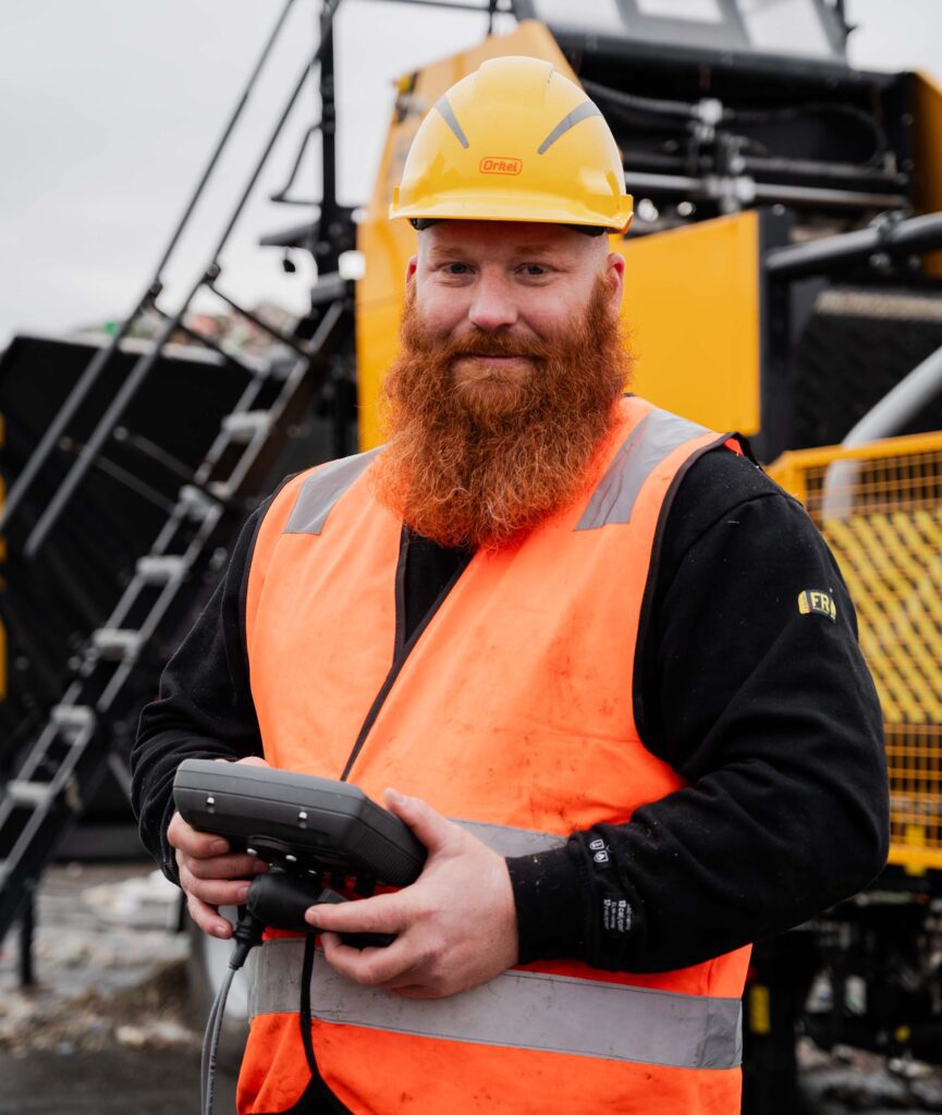 Un homme en gilet de sécurité et casque se tient avec confiance dans une installation de déchets, avec une grande machine industrielle en arrière-plan. Le cadre inclut des piles de matériaux recyclables et des équipements lourds, soulignant un environnement industriel axé sur la gestion et le traitement des déchets.