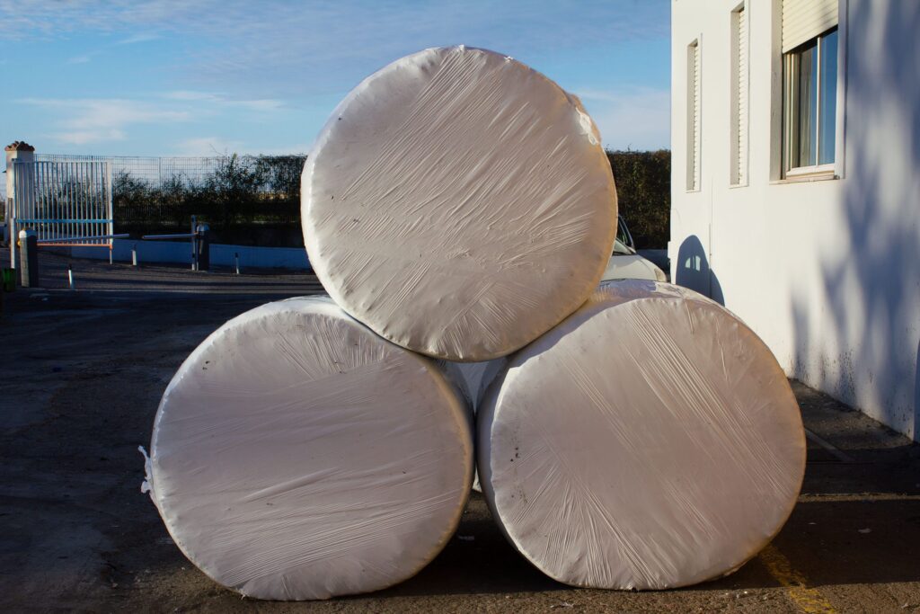 Trois balles rondes emballées dans du plastique blanc, empilées soigneusement à côté d'un bâtiment blanc.
