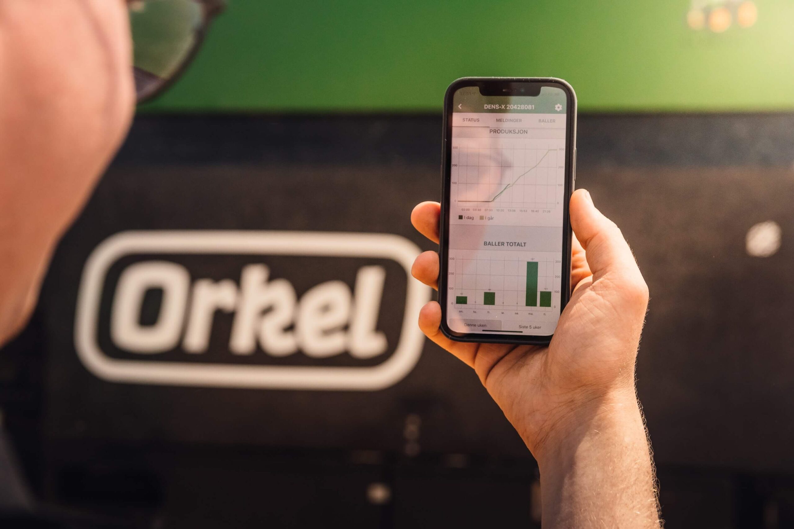 A man in the foreground holding up a smartphone. The phone is in focus showing an app. An agricultural baler is visible in the background.