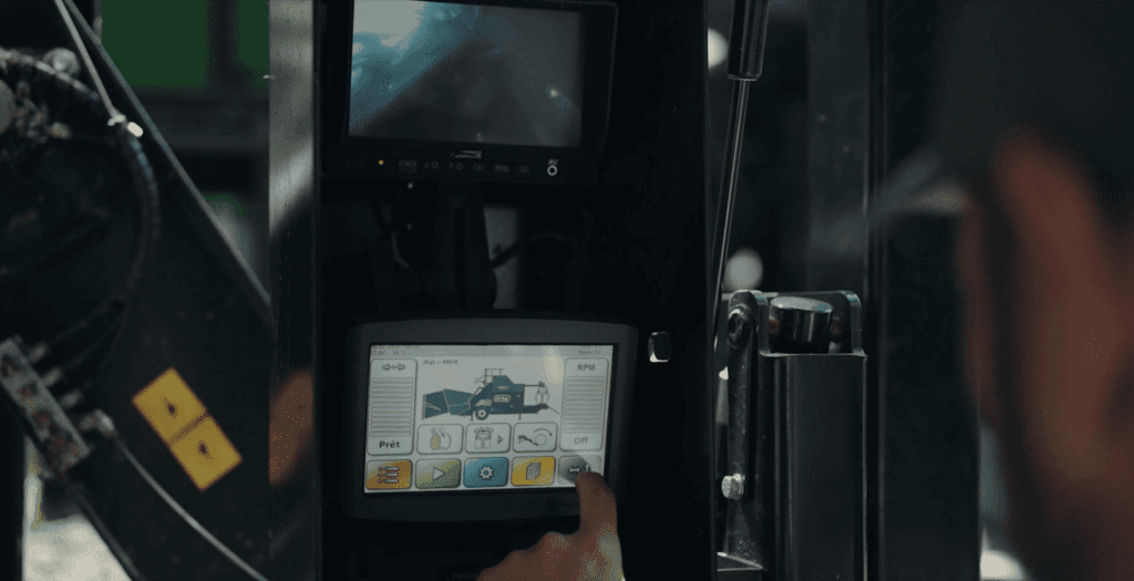 Nahaufnahme eines Touchscreens, der zur Bedienung einer landwirtschaftlichen Presse verwendet wird. Im Vordergrund sehen wir verschwommene Konturen eines Mannes, der den Touchscreen bedient. 