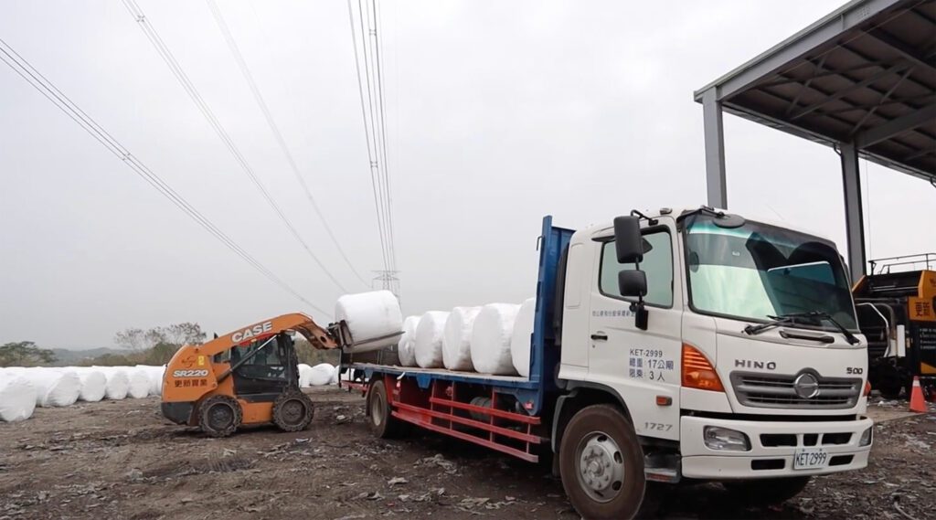 A forklift loading round bales onto a truck for transport, outside a large shed