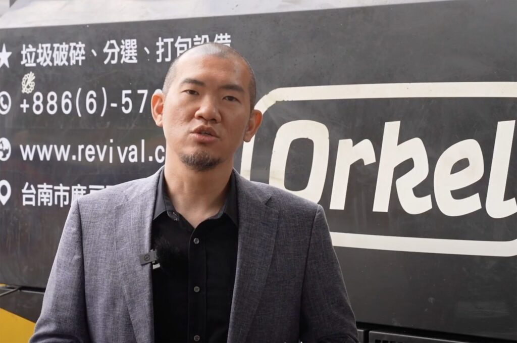 A man being interviewed, wearing a suit and a black polo shirt. An industrial machine in the background